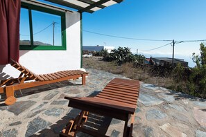 A patio with two wooden lounge chairs and a table, a window with a green frame, and a view of the landscape. - Casas Rurales Herreñas (Valverde)