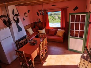 A cozy room with a wooden sofa, a dining table with chairs, a refrigerator, and a window with curtains.