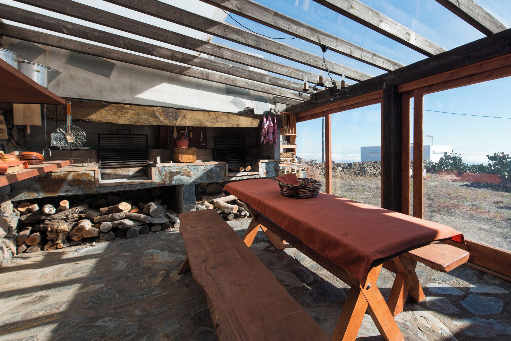 Cocina rústica al aire libre con un hogar de piedra, bancos de madera y vistas amplias por ventanas grandes.