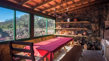 A rustic cabin interior with a wooden table, benches, shelves with kitchenware, and a stone wall.