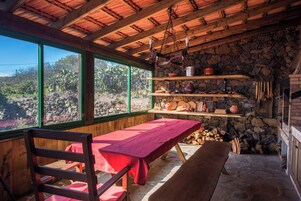 A rustic cabin interior with a wooden table, benches, shelves with kitchenware, and a stone wall.