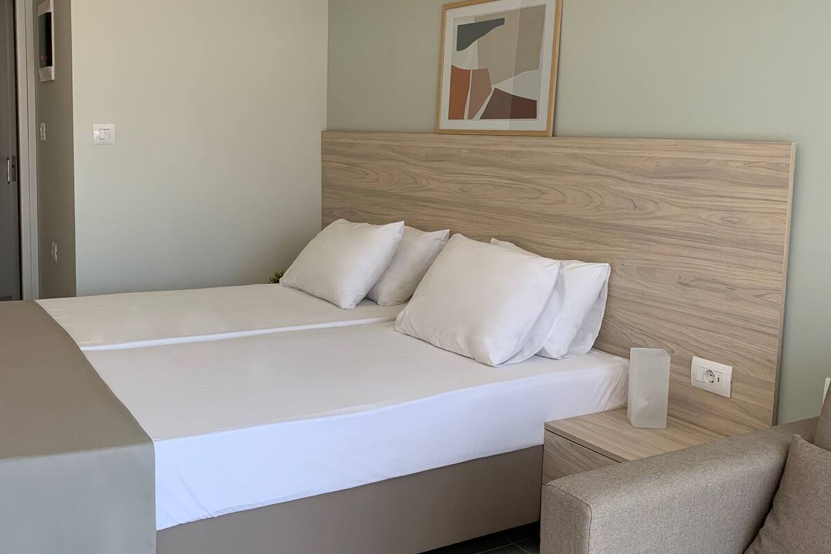 A modern hotel room with a wooden headboard, a large bed with white linens, a bedside table with a lamp, and a framed abstract painting on the wall.