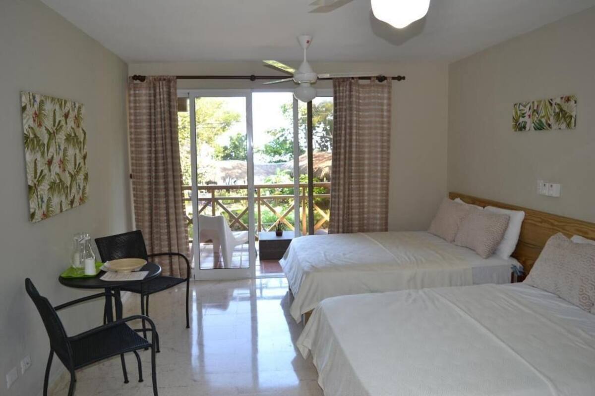 A hotel room with two beds, a dining table with chairs, a ceiling fan, and a balcony with curtains.