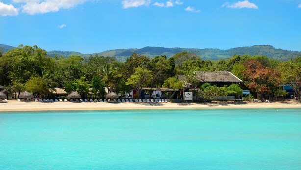 A beachside resort with thatched-roof huts, a sandy beach, and a backdrop of green hills. - Paradise Island Beach Resort (Punta Rucia)