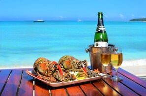 A lobster on a plate, a bottle of champagne, and two glasses on a wooden table with a beach and ocean in the background.