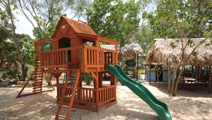 A wooden play structure with a slide and climbing area, surrounded by trees and a thatched-roof building. - Paradise Island Beach Resort (Punta Rucia)