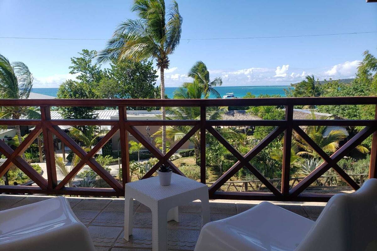 A balcony with a view of the ocean, palm trees, and a small table.