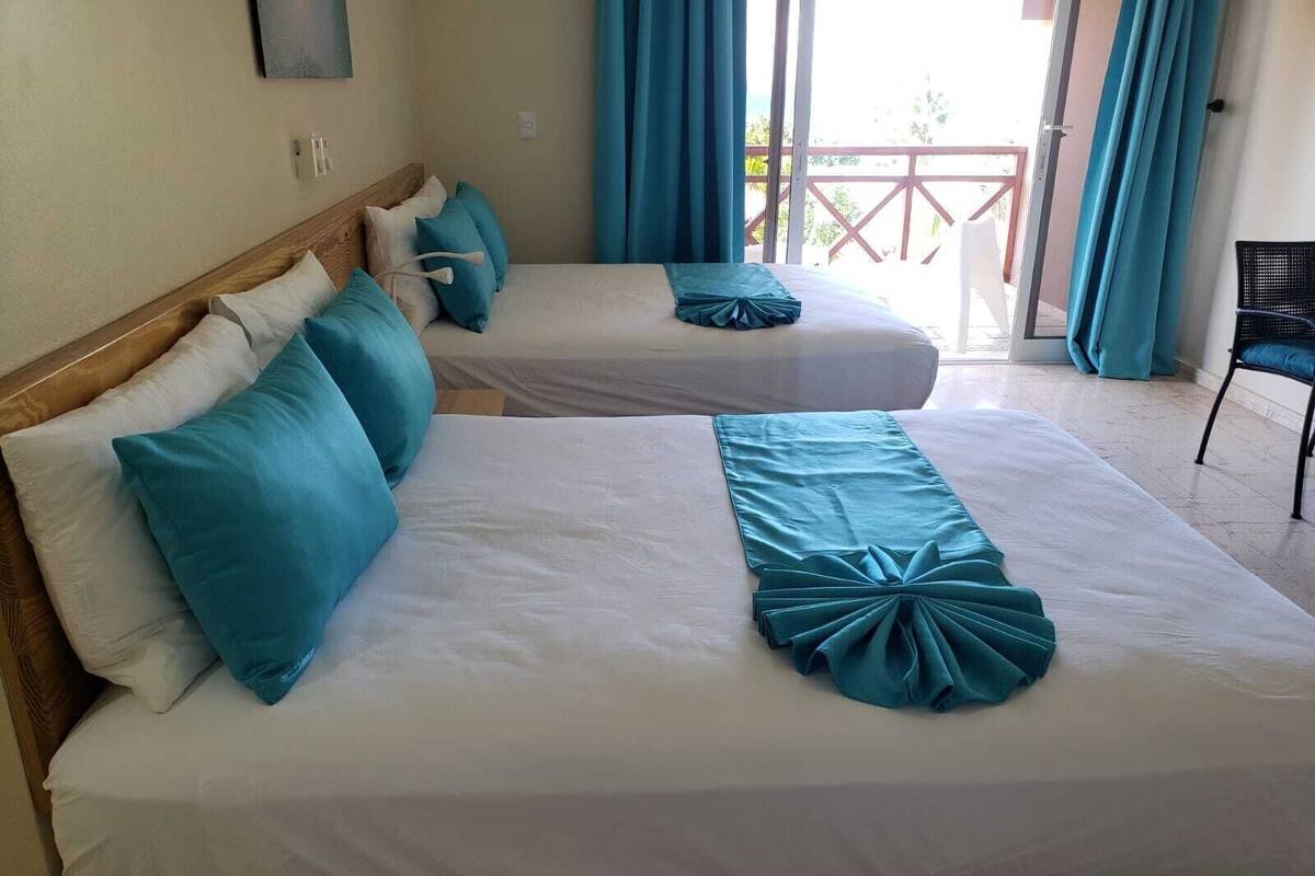 A hotel room with two beds, each with white linens and teal accents, a wooden headboard, and a view of the outdoors through a glass door.