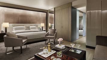 Quarto de hotel moderno com uma cama grande, duas poltronas, uma mesa de centro com amenidades e vista para o banheiro.