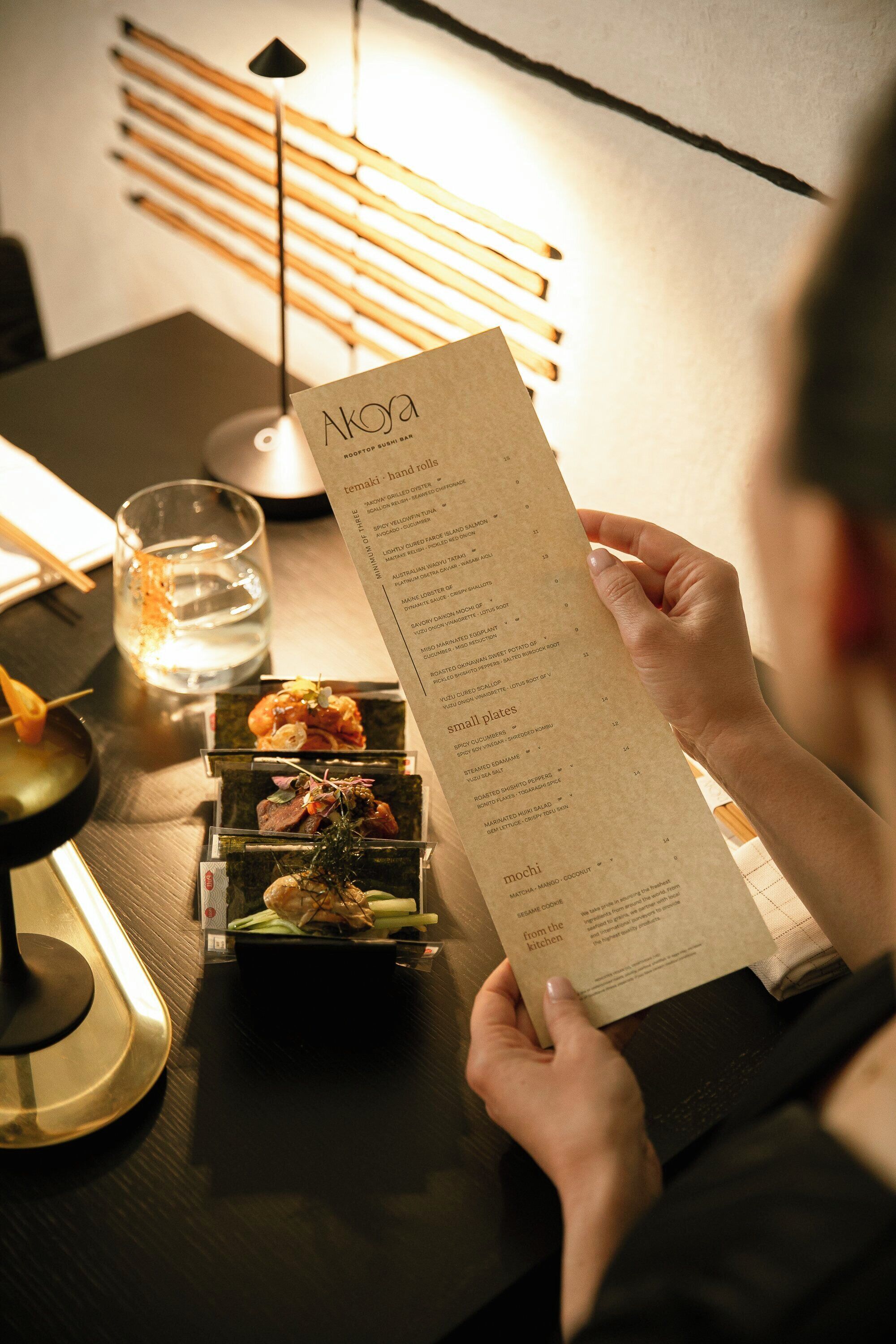 A person holding a menu with various dishes listed, a glass of water, and a candle on a table.
