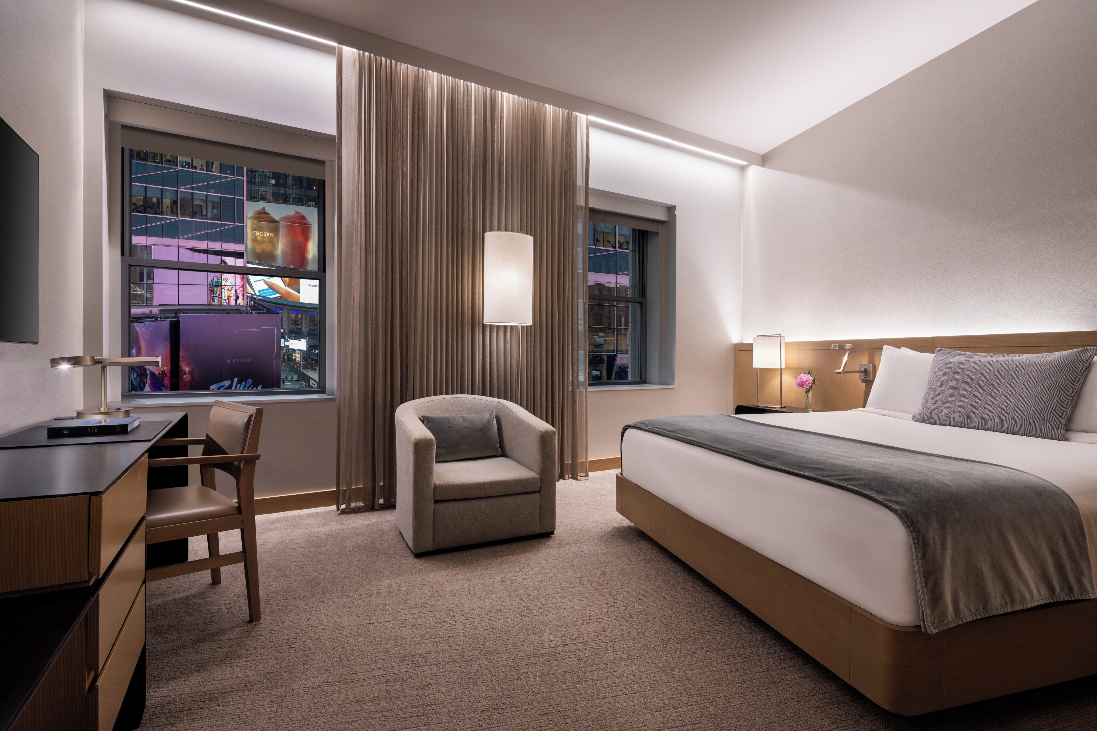 Una habitación de hotel con cama, escritorio, silla y un gran ventanal con cortinas.