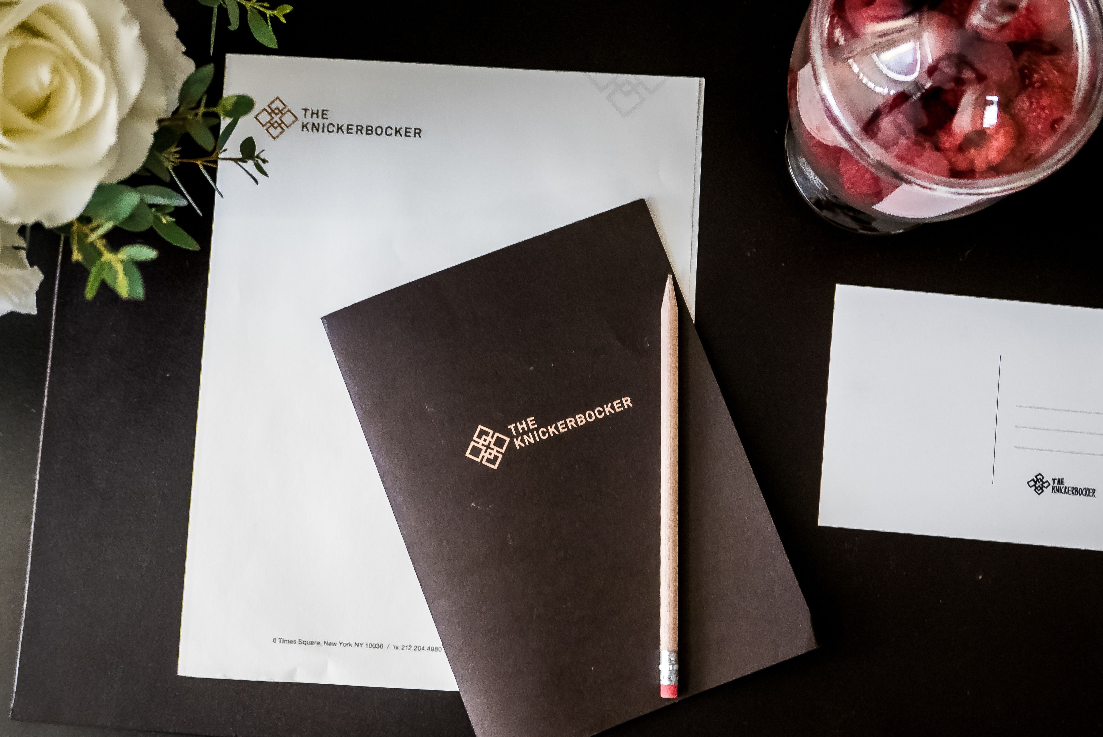 A black notebook with 'The Knickerbocker' logo, a pencil, a white envelope, and a jar of raspberries on a dark surface.