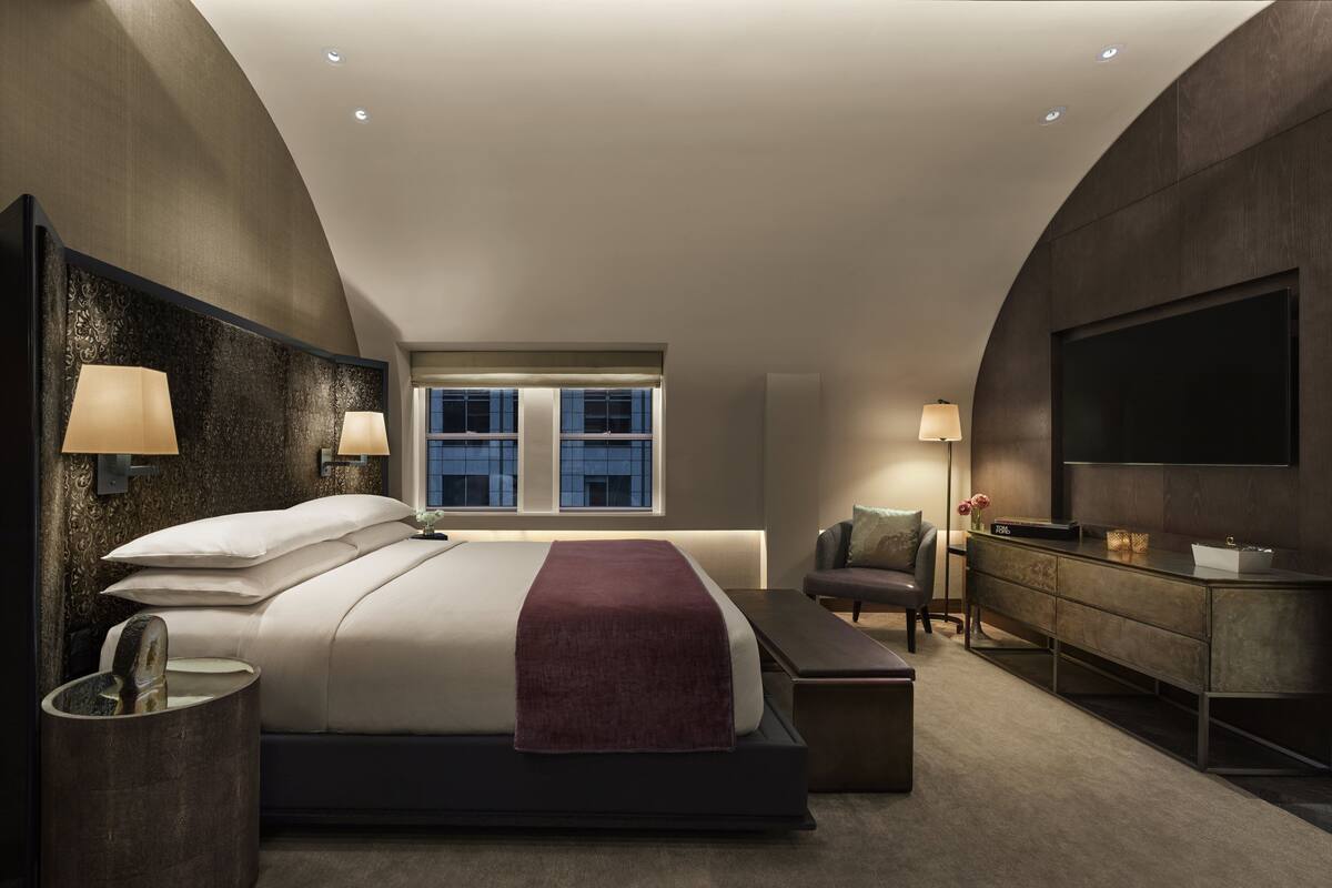Un dormitorio moderno con una cama grande, lámparas de noche, un televisor, un aparador y una ventana con vistas a la ciudad.