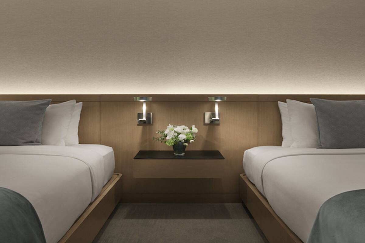 Habitación de hotel con dos camas, una mesita de noche con un jarrón de flores e iluminación en la pared.