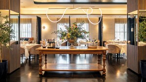 A restaurant with a central wooden table, multiple dining tables, and chairs. There is a large vase with flowers on the central table.