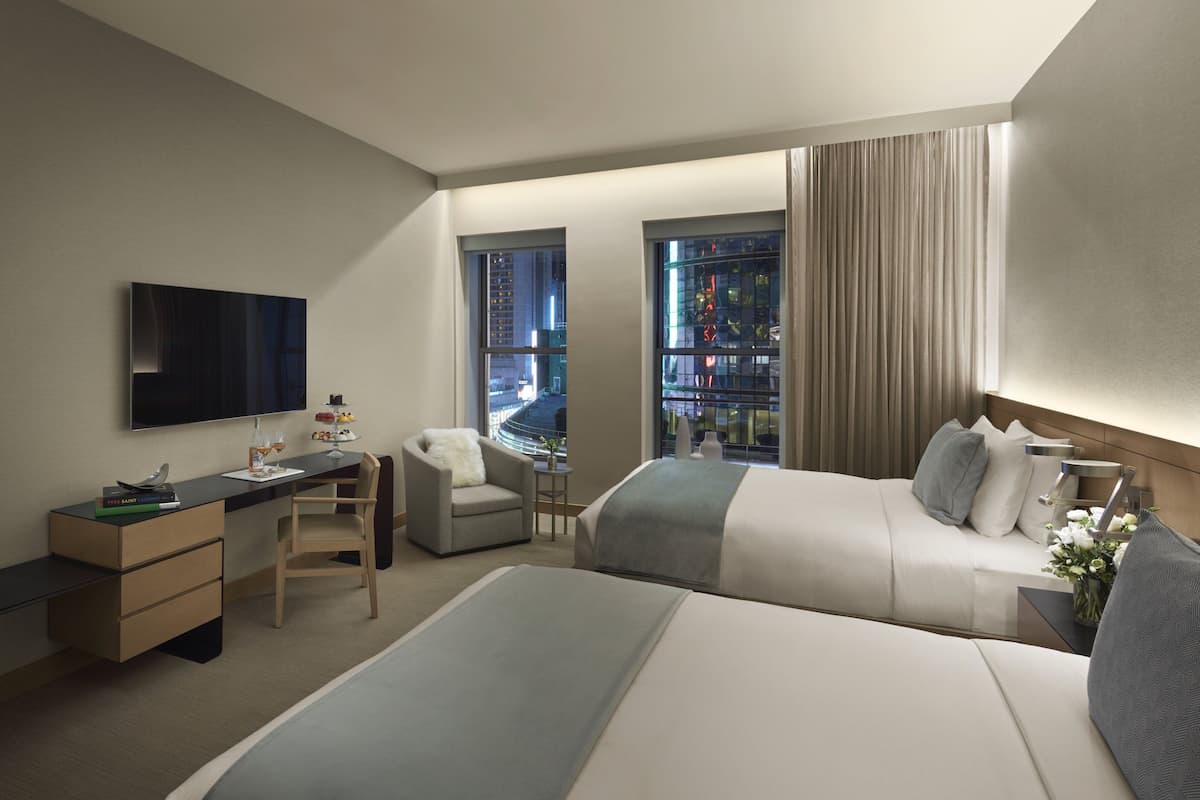 Una habitación de hotel moderna con una cama grande, un escritorio con televisión de pantalla plana, una silla y vistas de la ciudad a través de amplios ventanales.