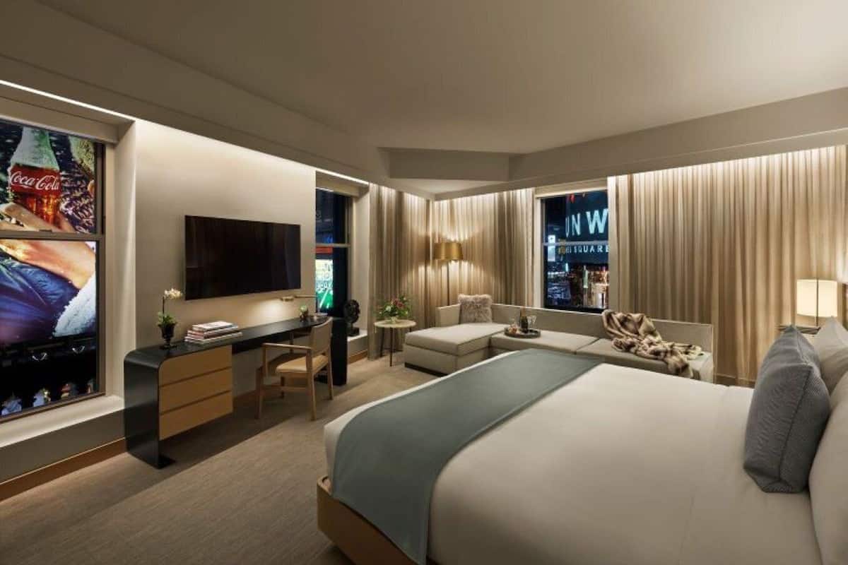 Una habitación de hotel moderna con una cama grande, un escritorio con televisor de pantalla plana, una zona de estar con sofá y sillones, y vistas a la ciudad desde la ventana.