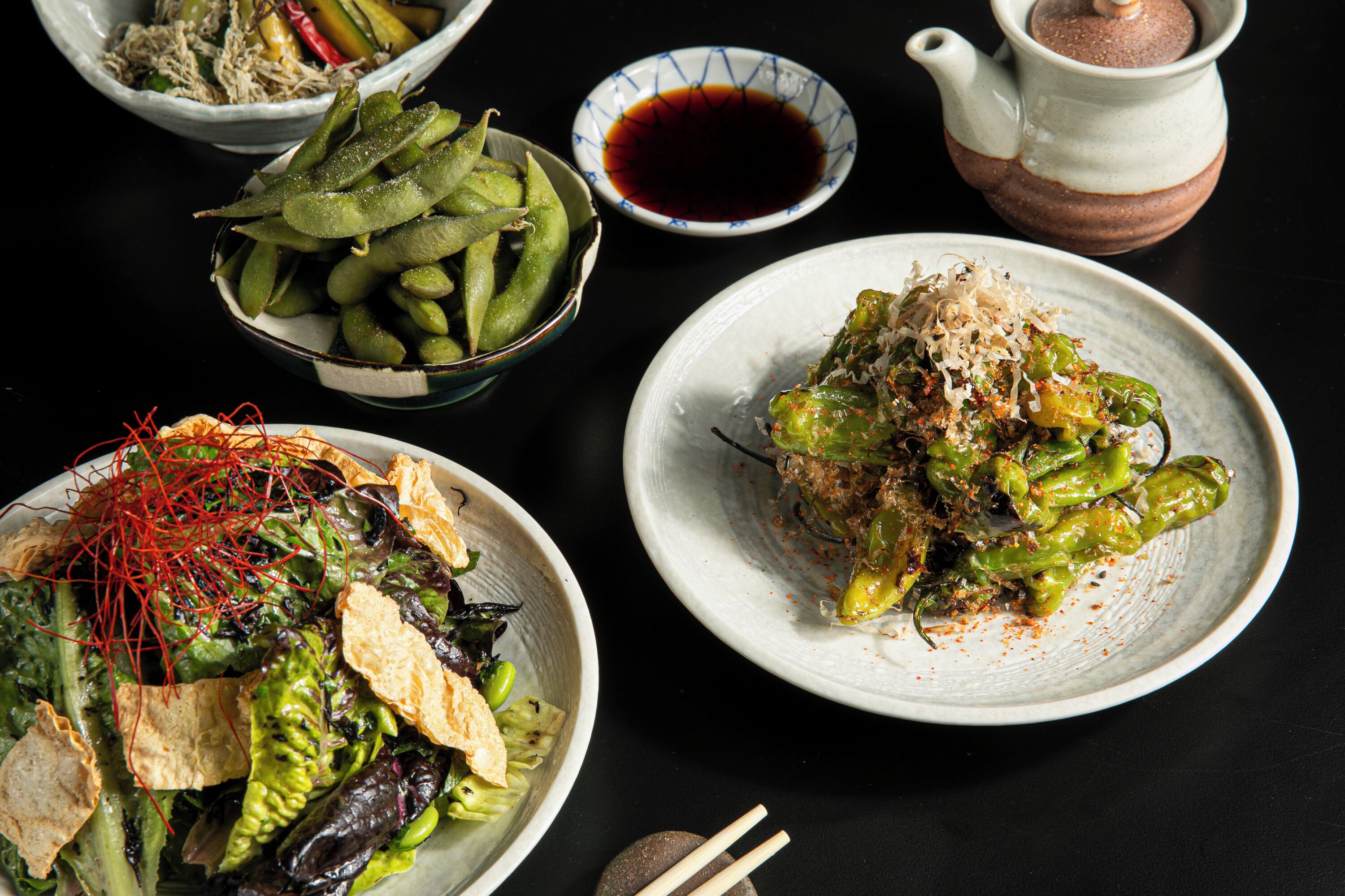 A table with various dishes, including a bowl of edamame, a plate of salad, and a bowl of dipping sauce.