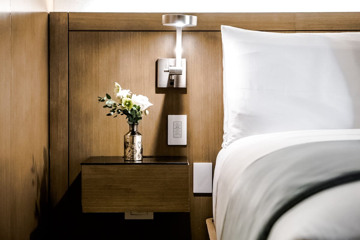 Una cama bien tendida con un cojín blanco y un cabecero de madera. Se observa una mesita de noche con un florero y una lámpara de pared.