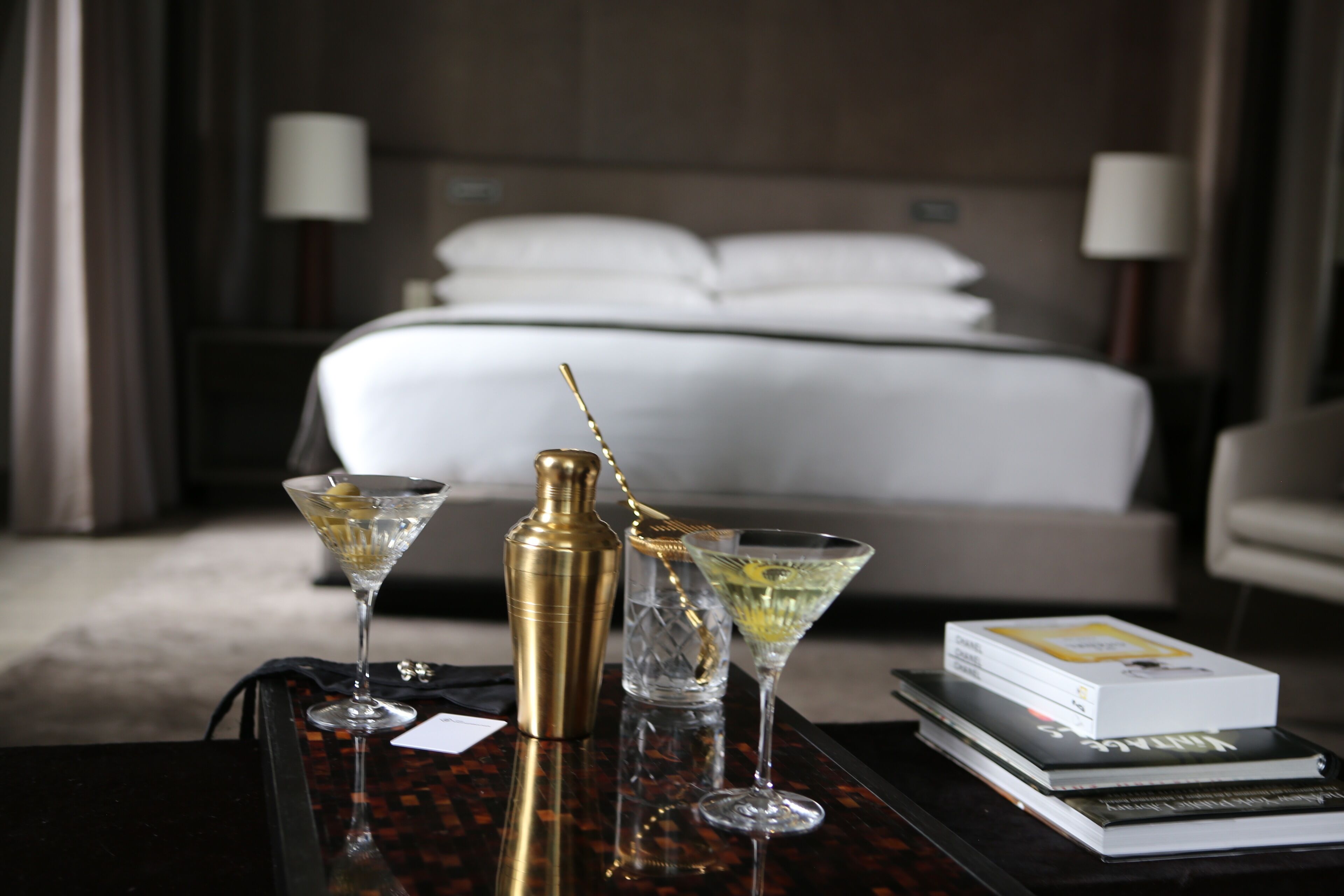 Une chambre d’hôtel bien rangée, avec un lit, une table de chevet sur laquelle se trouvent un verre à martini, un shaker à cocktails et un livre.