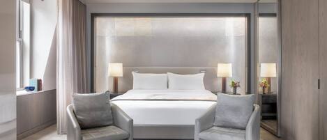 A modern hotel room with a large bed, two armchairs, a coffee table with a book, and a view of the city through the window.