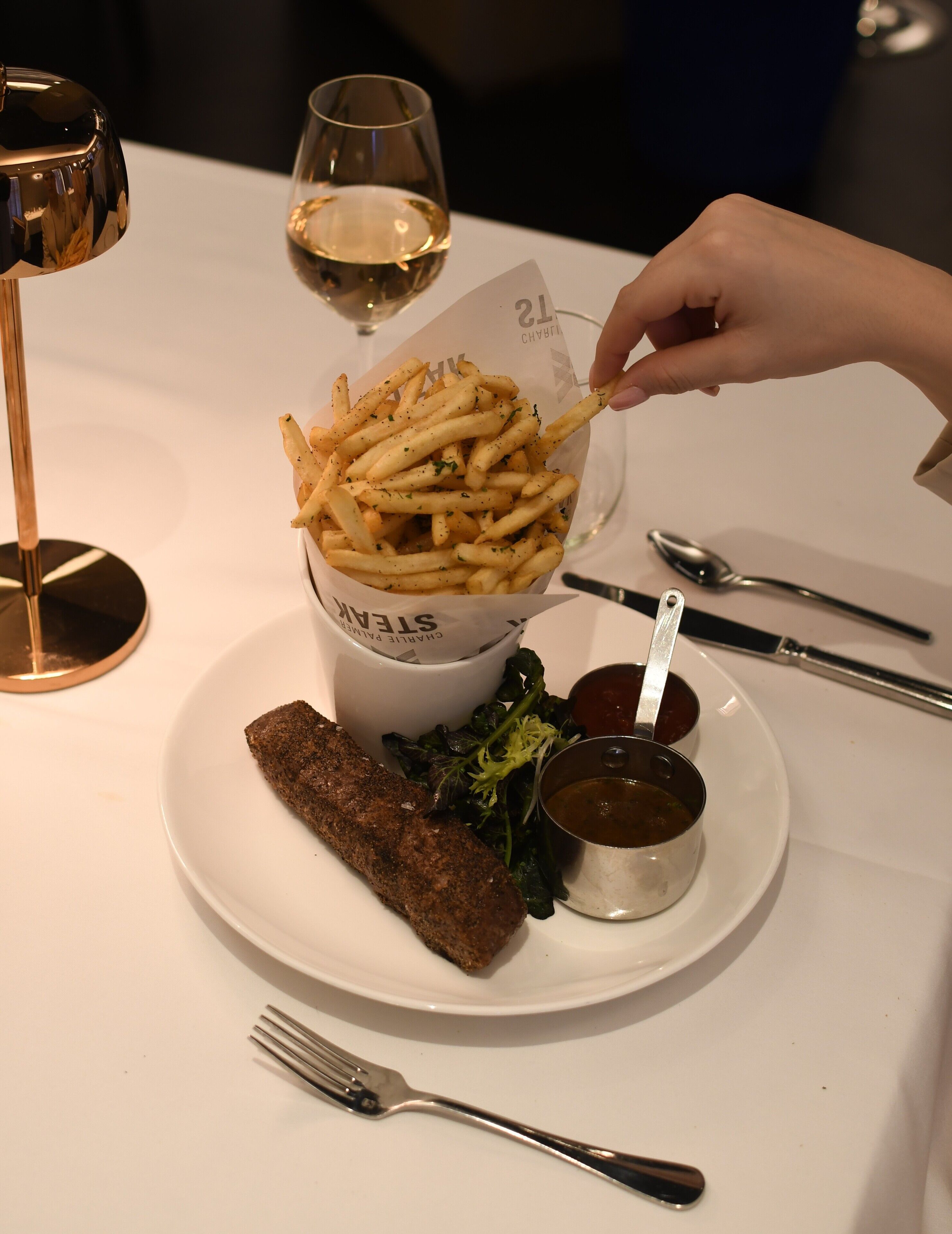 A plate with fries, a meat dish, and two sauces, with a fork and spoon on the table.