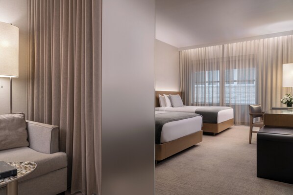 A hotel room with a sofa, two beds, a desk, and a large window with curtains.