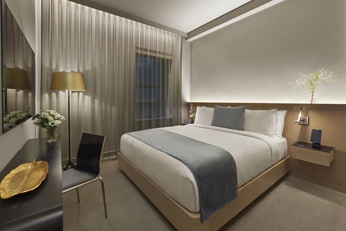 Una habitación de hotel moderna con una cama grande, un escritorio con una silla, una mesita de noche con lámpara y una ventana con cortinas.