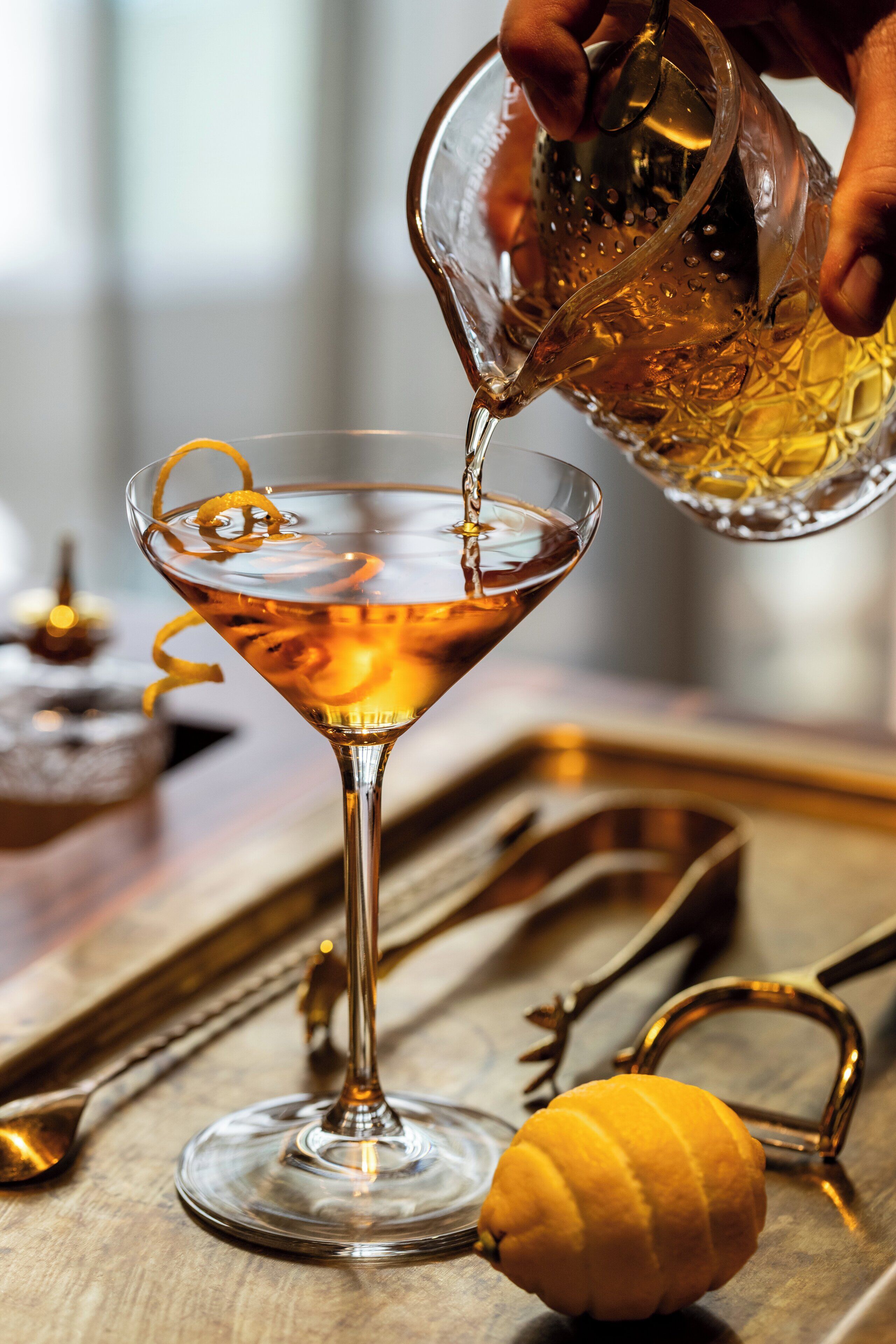 Een martini glas wordt gevuld met een gouden vloeistof, een citroen en een citroenschil als garnering op een houten ondergrond, met een lepel en een metalen ring.