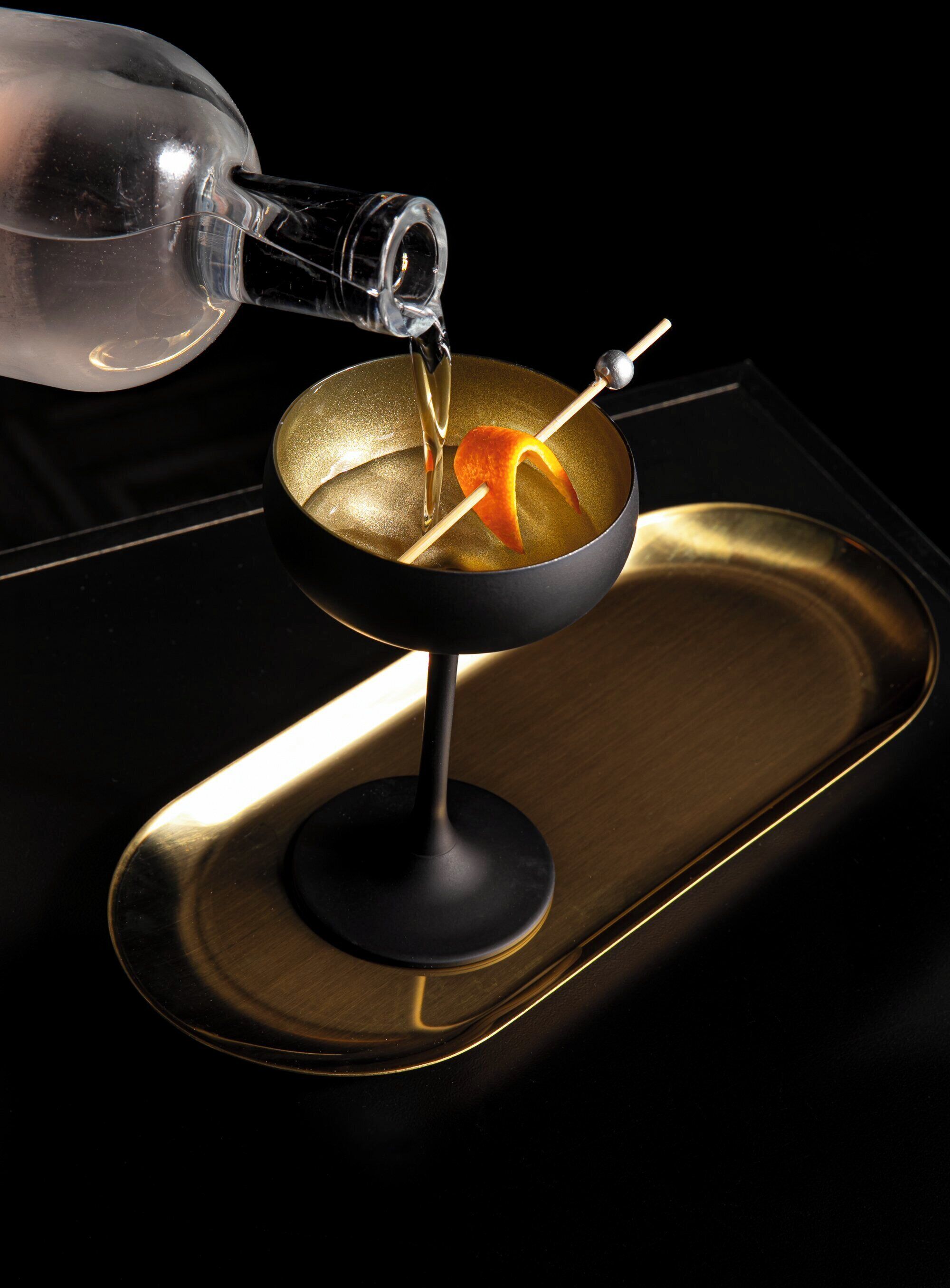 A cocktail being poured into a martini glass with an orange peel garnish on a black tray.