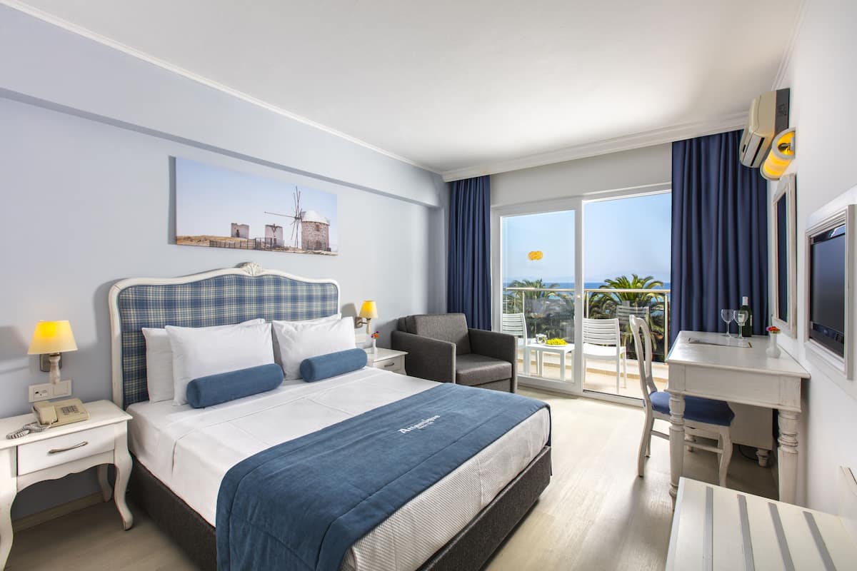 Une chambre d’hôtel avec un lit, un bureau, une chaise, un balcon avec vue et un tableau au mur.