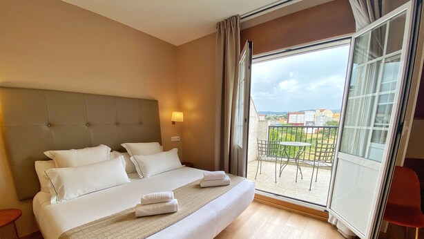 A hotel room with a large bed, a balcony with a table and chairs, and a view of buildings. - Hotel Ribeira Sacra (Monforte de Lemos)