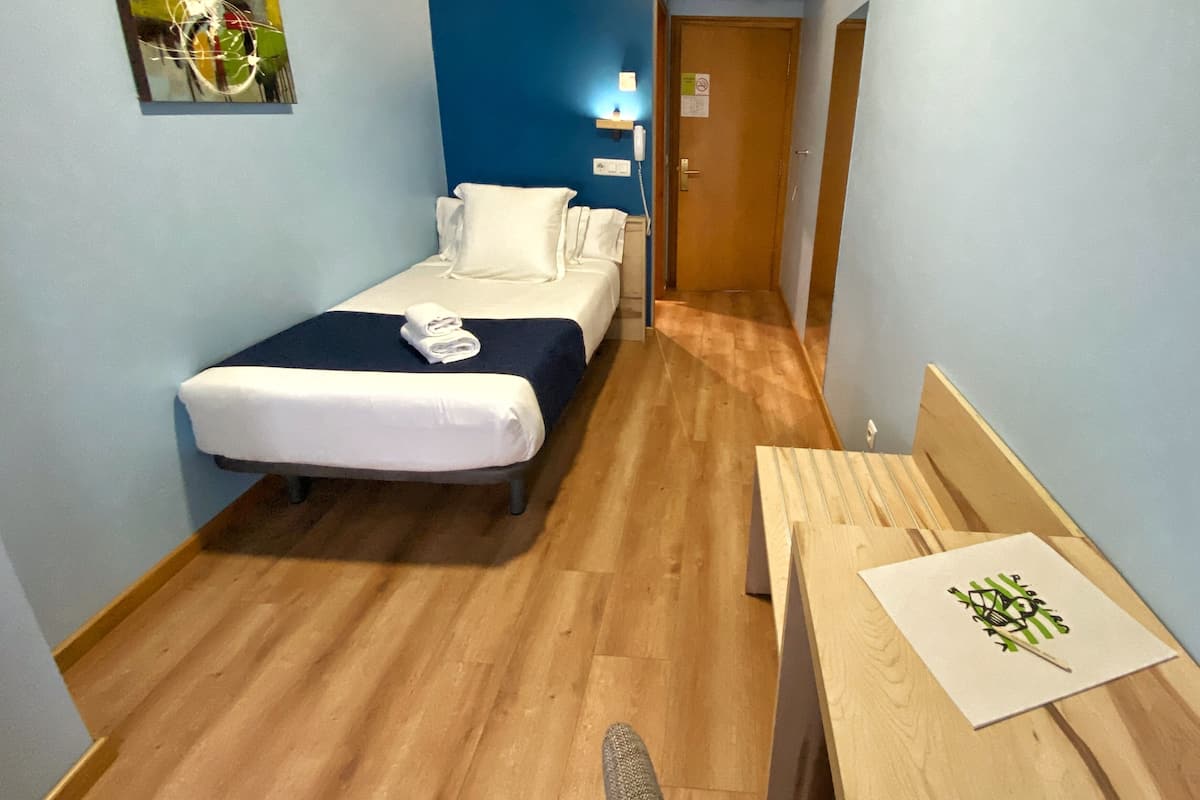 A small, narrow hotel room with a single bed, a bedside table, and a chair. There is a wooden floor, a blue wall, and a door leading to another room.