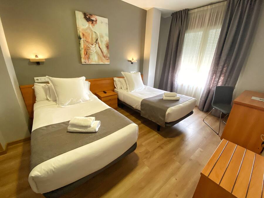 Ein Hotelzimmer mit zwei Betten, einem Schreibtisch, einem Stuhl, einem Bild an der Wand und einem Fenster mit Vorhängen.