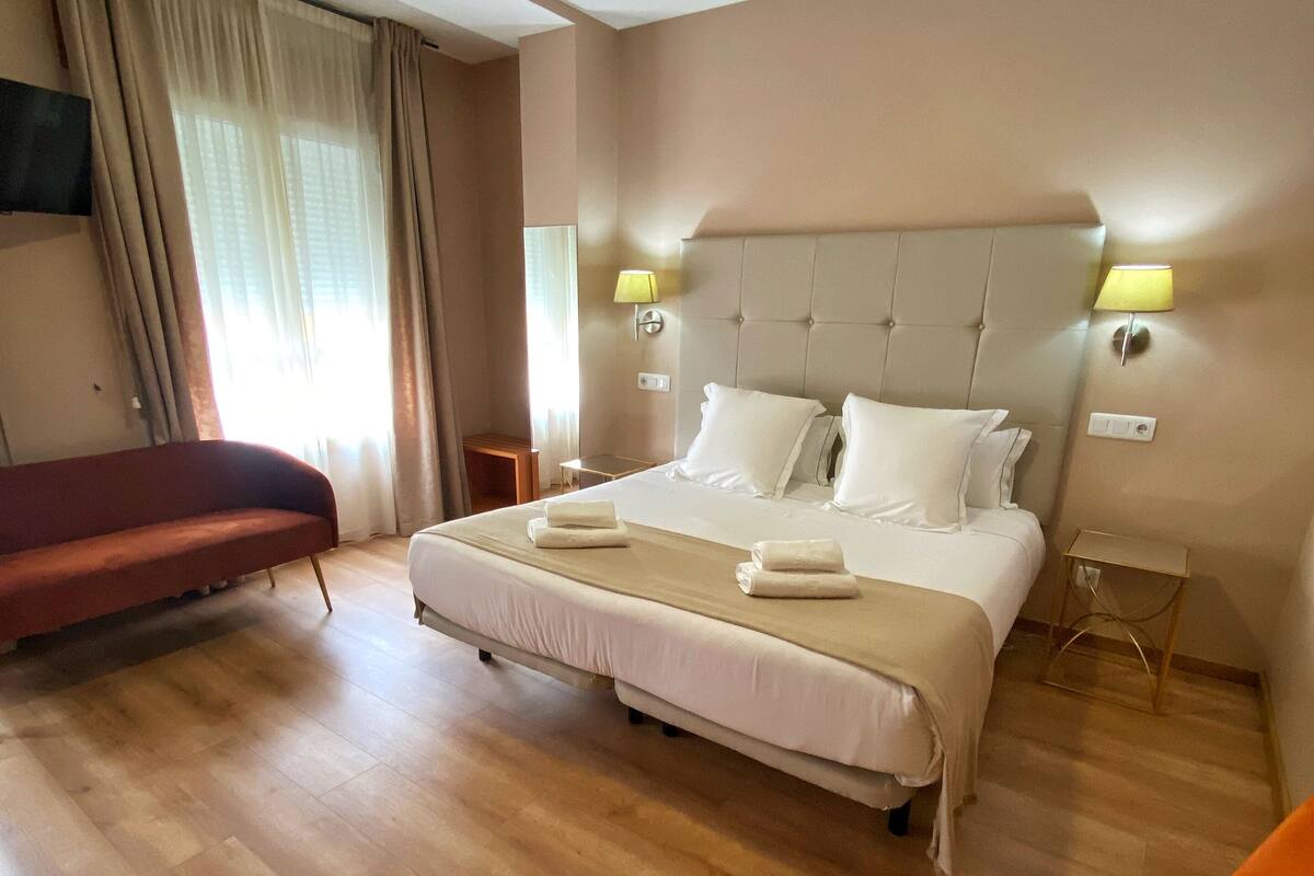 A hotel room with a large bed, a bedside table, a chair, and a television on the wall.