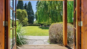 Uma porta de madeira aberta dá para um jardim com gramado, árvores e um caminho de cascalho.