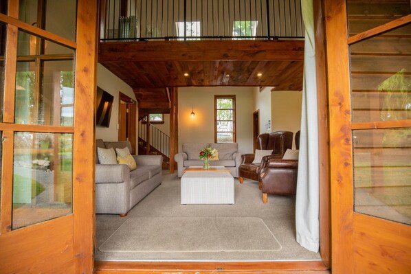 A cozy living room with a sofa, armchair, and coffee table, wooden beams on the ceiling, and large windows.