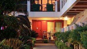 Un hotel con puerta de entrada roja, un balcón con barandilla y un camino bordeado de vegetación.