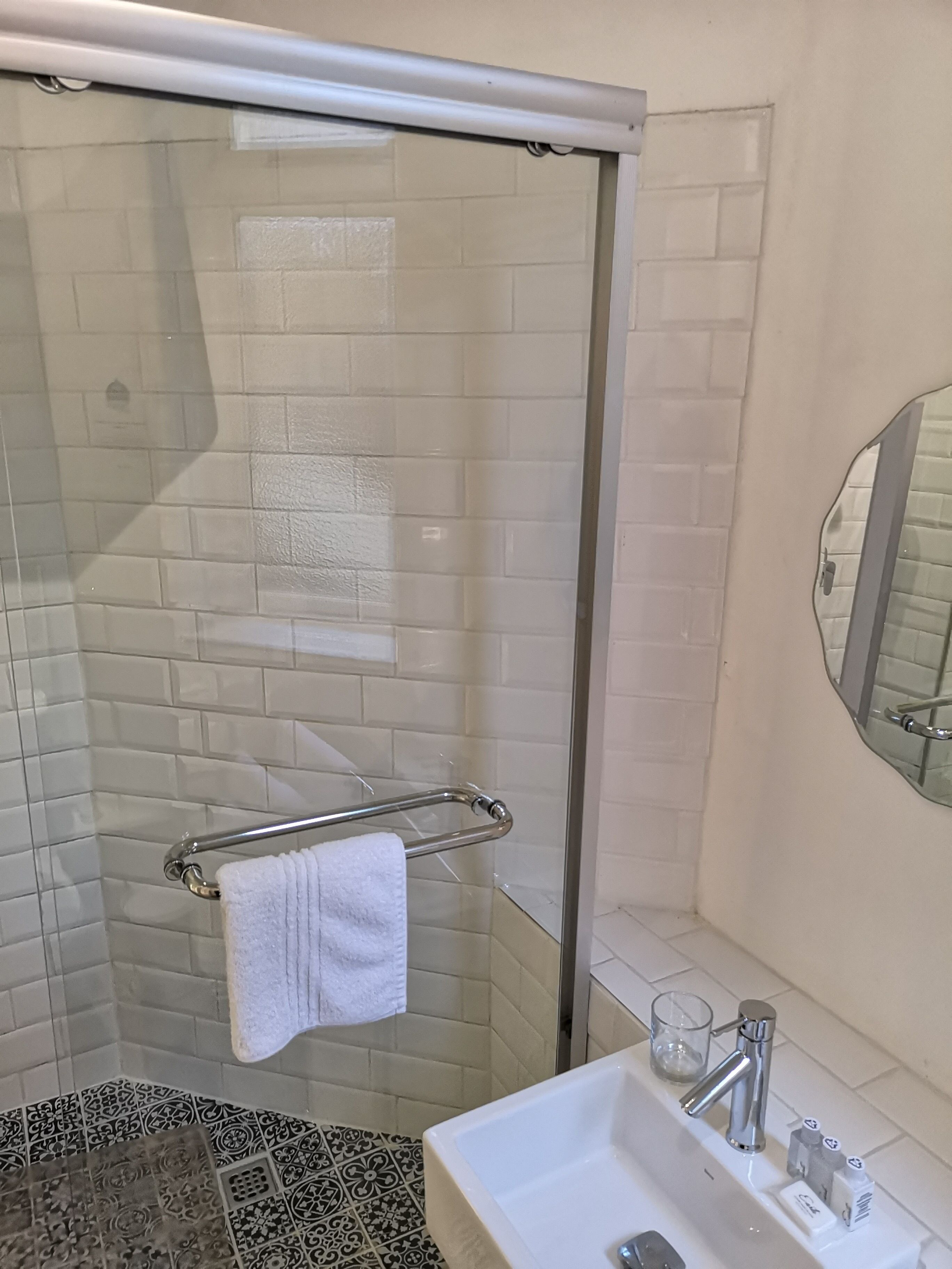 A bathroom with a glass shower enclosure, a white sink with a mirror, and patterned tiles on the floor.