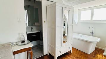 A spacious bathroom with a wooden floor, a white bathtub, a sink, and a vanity with a mirror. There are framed artworks on the wall and a door leading to another room.