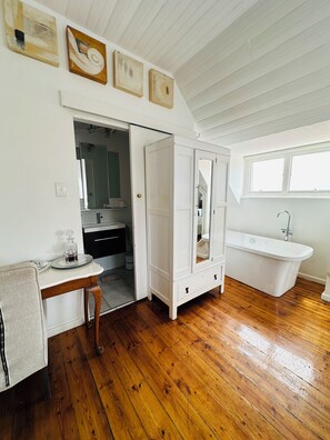 A spacious bathroom with a wooden floor, a white bathtub, a sink, and a vanity with a mirror. There are framed artworks on the wall and a door leading to another room.