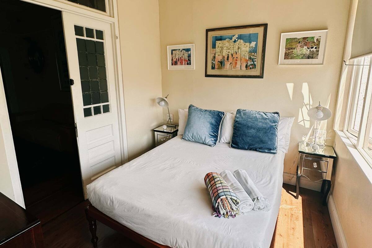 A bedroom with a wooden bed, white bedding, a chandelier, a nightstand, and a window with blinds.