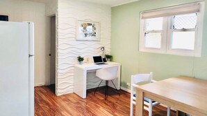 A small, well-lit room with a wooden floor, a white desk with a laptop, a chair, and a dining table. There is a window with blinds and a door to the left.