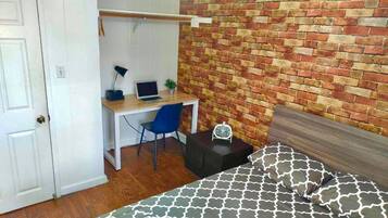 A bedroom with a bed, a desk with a laptop, a chair, and a brick-patterned wall.