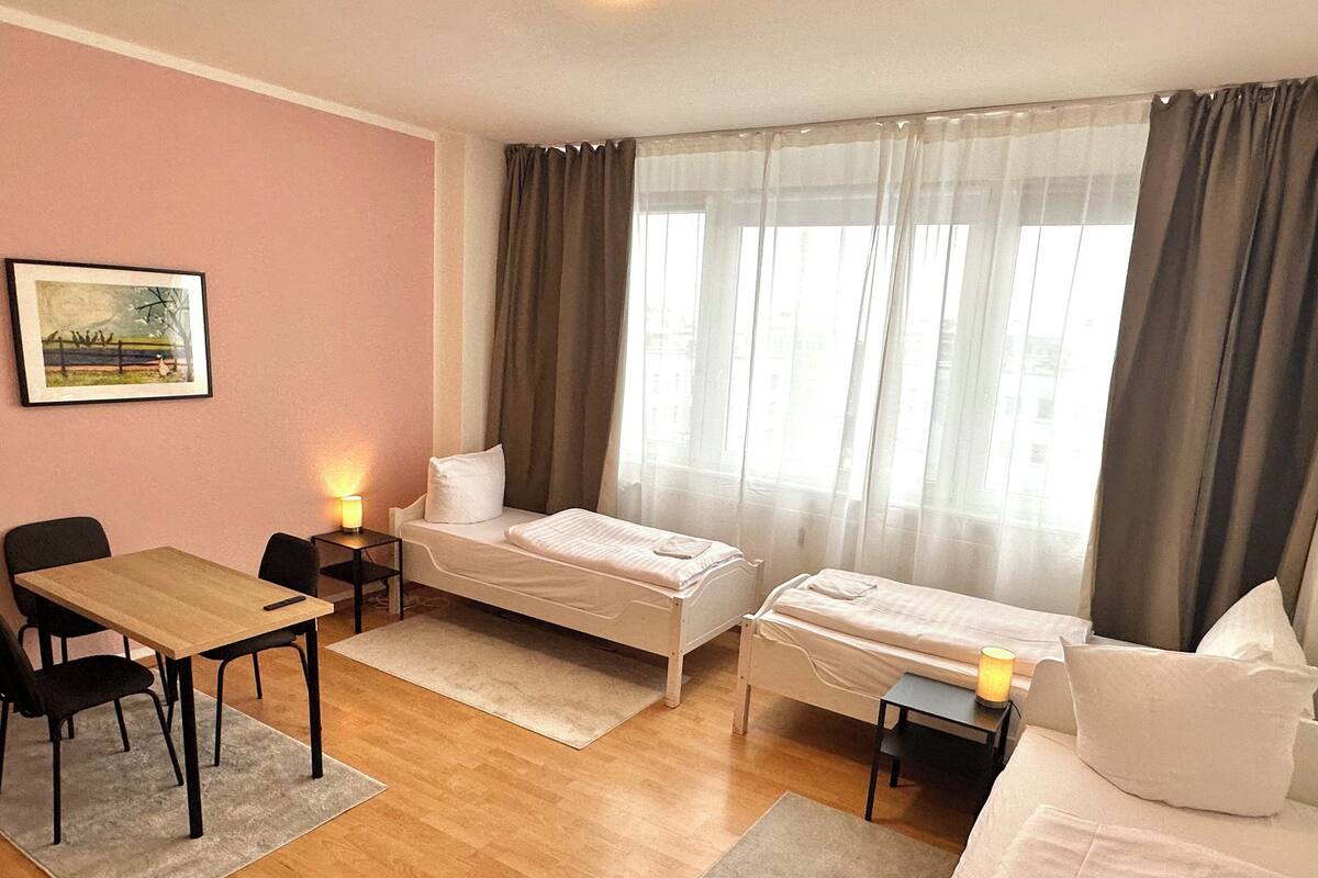 A hotel room with two single beds, a wooden table with chairs, a small desk, and a painting on the wall.