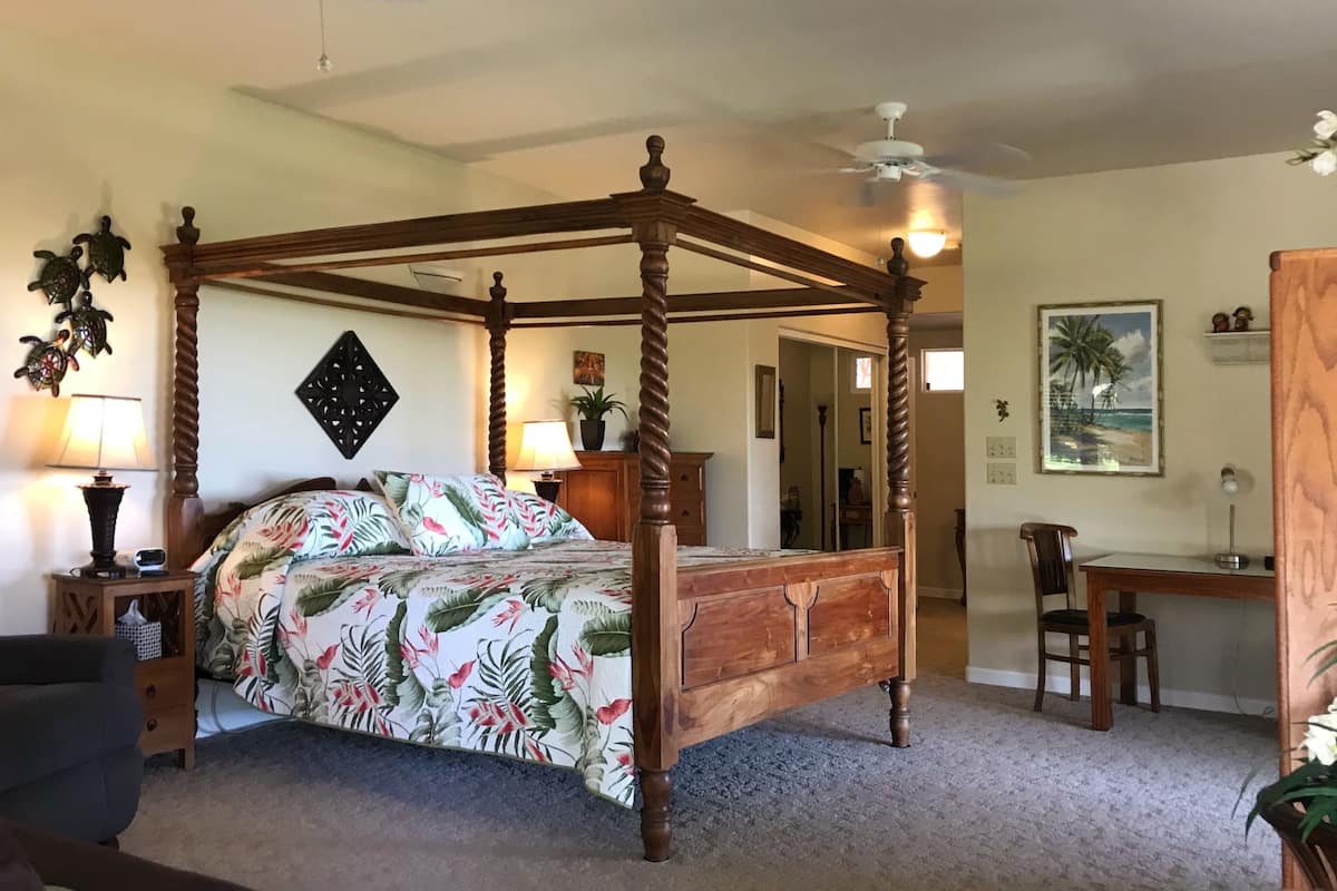 A four-poster bed with floral bedding, a desk with a chair, a ceiling fan, and a painting of a beach scene.