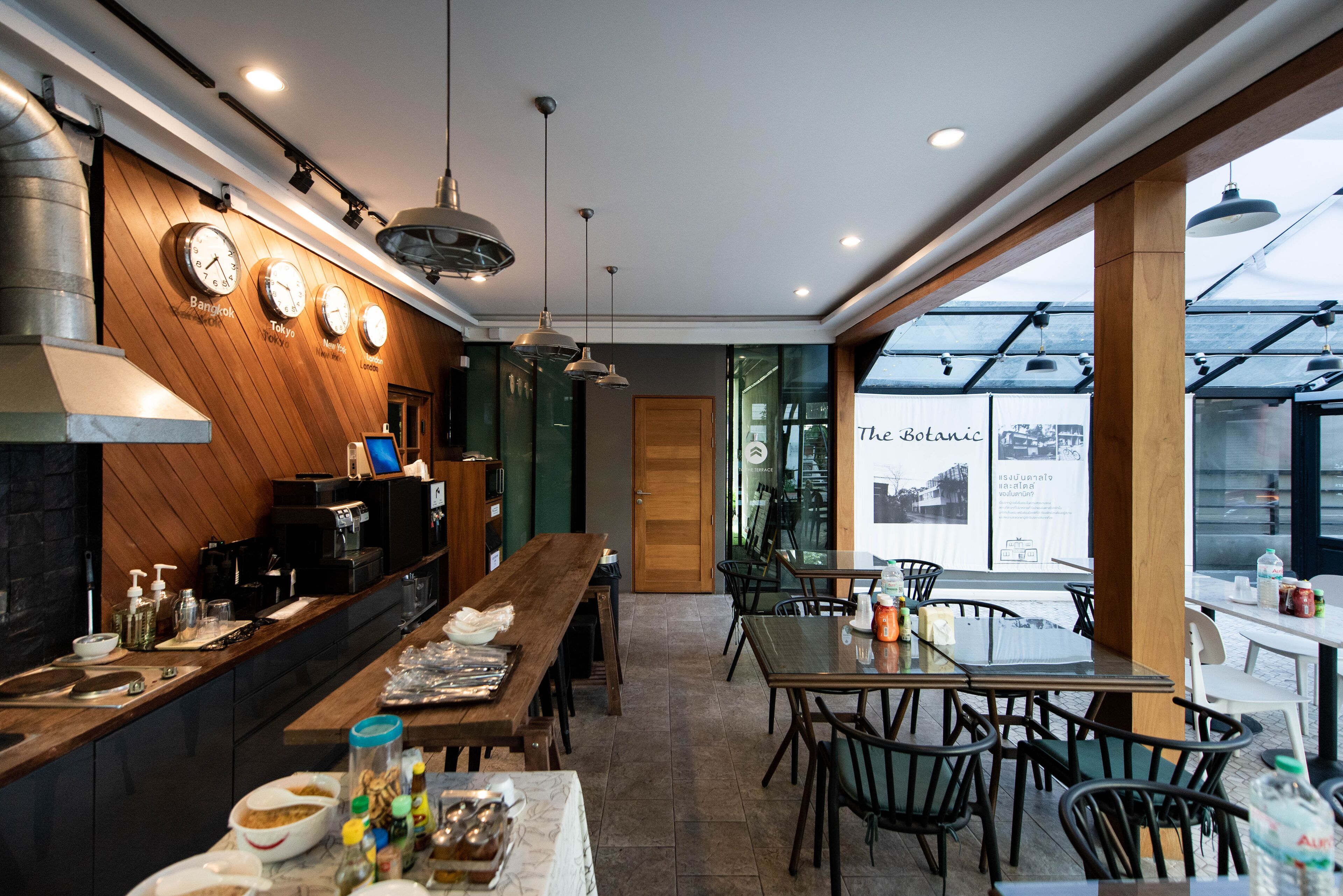 A modern restaurant with a long wooden counter, multiple dining tables, and chairs. There are clocks on the wall and a large window with a view.