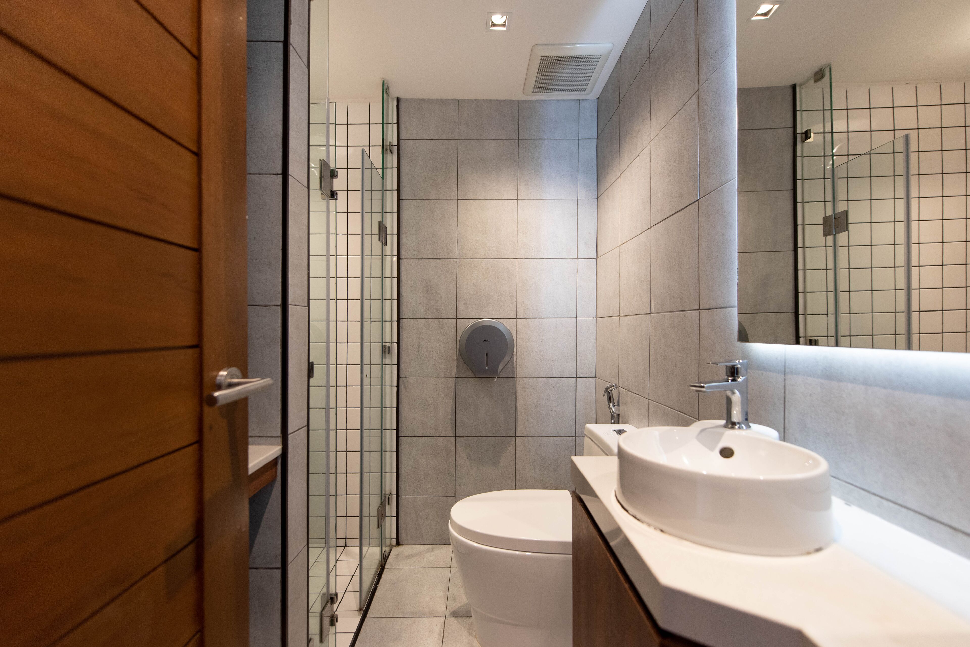 Un baño moderno con puerta de madera, cabina de ducha de cristal, inodoro blanco y lavamanos con espejo superior.