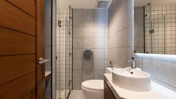 Un baño moderno con puerta de madera, cabina de ducha de cristal, inodoro blanco y lavamanos con espejo superior.
