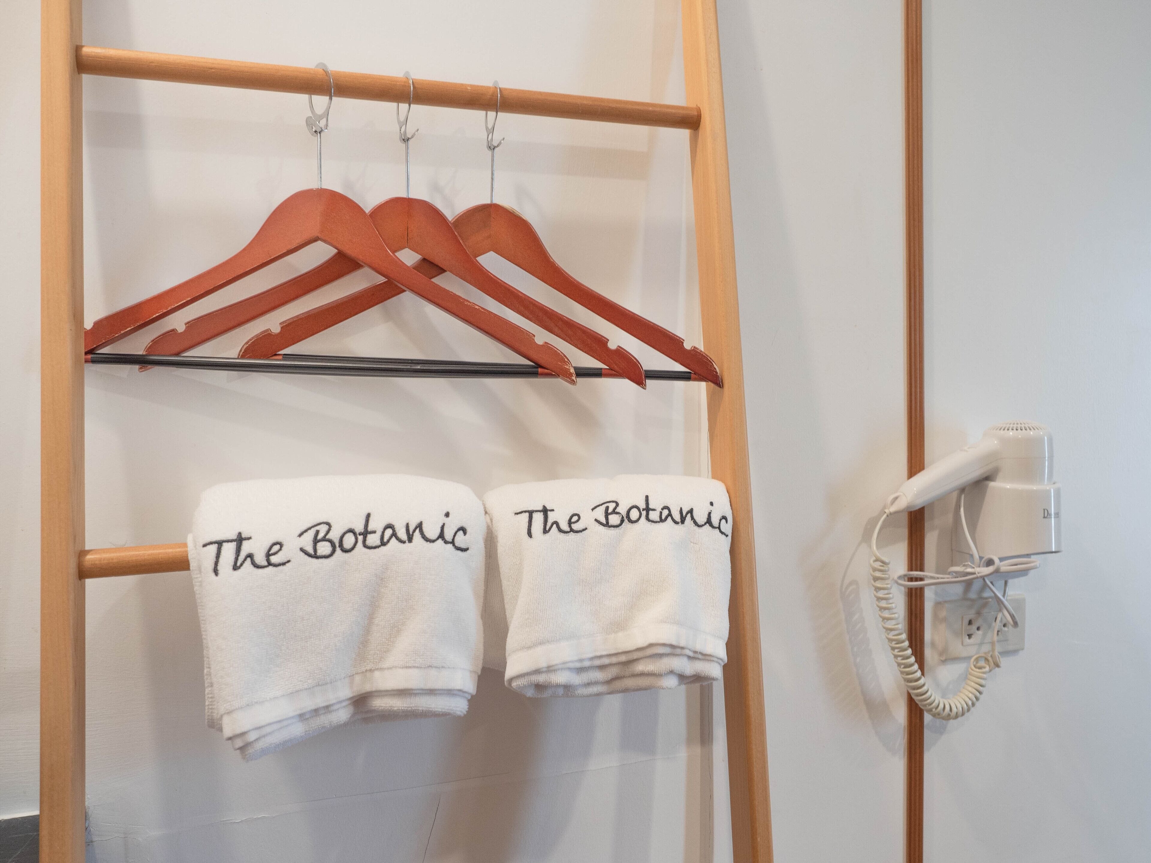 Un perchero de madera con dos toallas blancas colgando, cada una con la etiqueta "The Botanic". Un secador de pelo blanco está montado en la pared a la derecha.